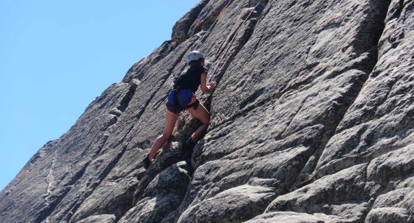 An outward bound student wearing safety gear climbs a rock wall under blue skies